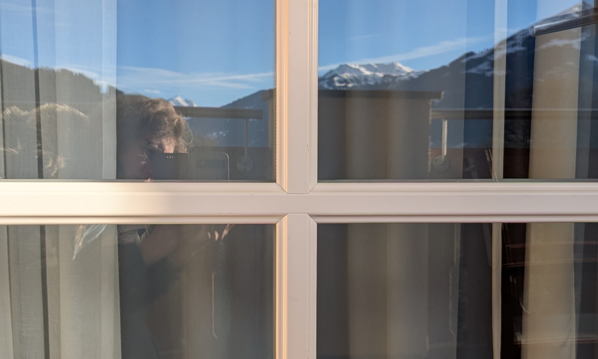 Eine Spiegelung der Alpen und von Dorothee, wie sie die Spiegelung fotografiert auf einer Balkontür