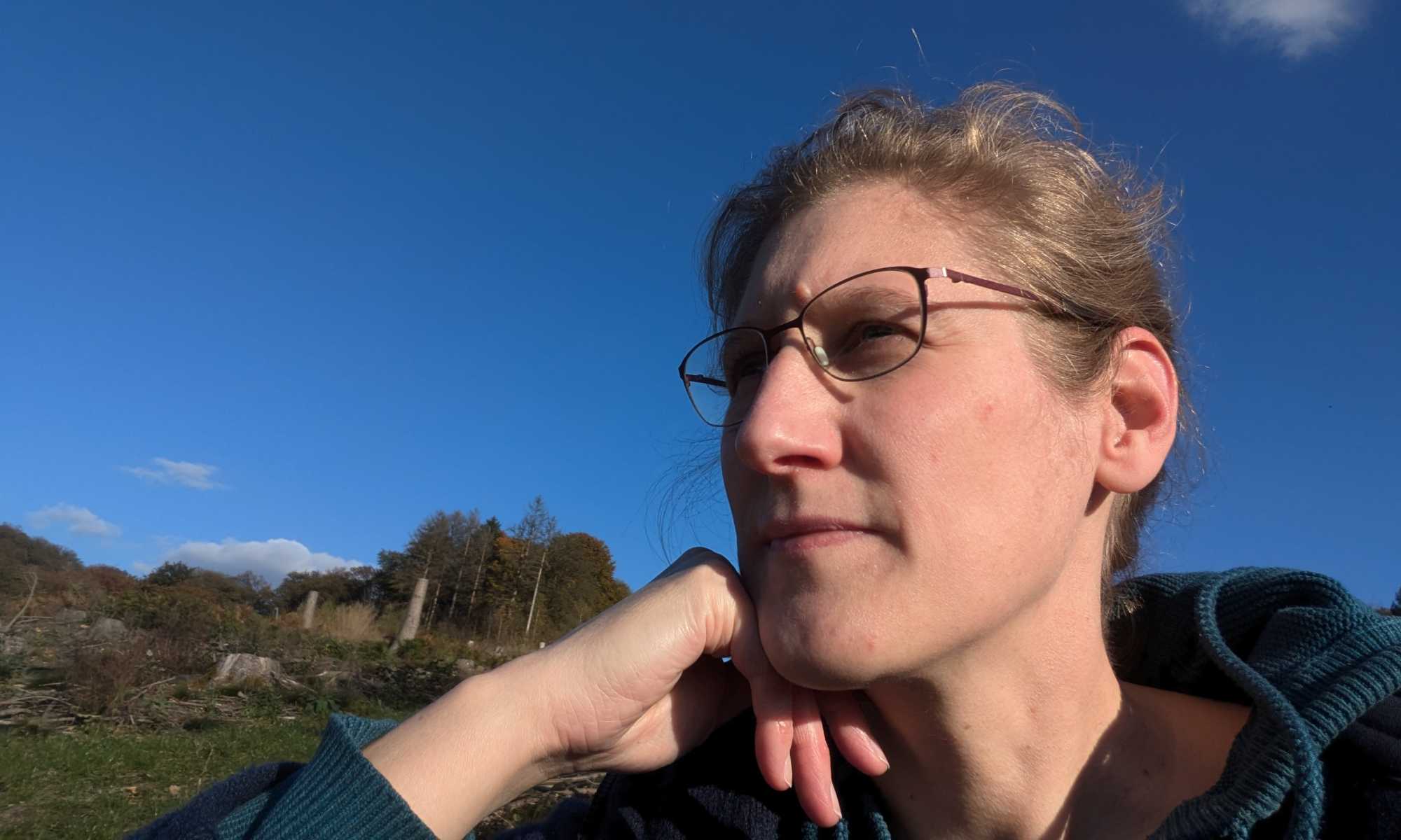 Dorothee sitzt mit dem Kopf auf die Hand gestützt vor blauem Himmel und Wald im Hintergrund