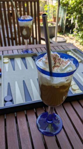 Auf dem Terrassentisch liegt ein aufgebautes Backgammonspiel, davor steht ein Eiskakao in einem hohen Glas mit einem Strohhalm aus Metall. Mittagspause deluxe
