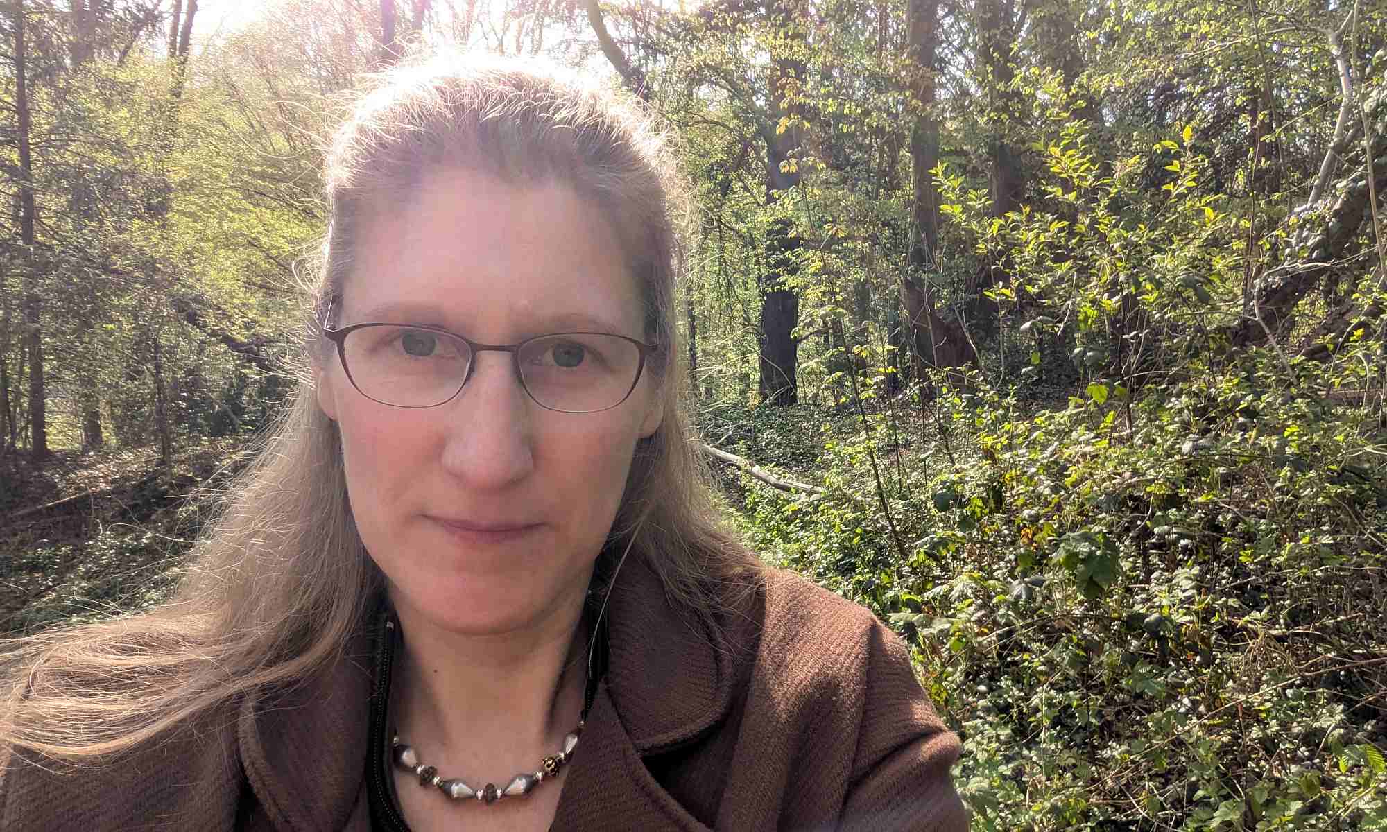 Dorothee steht im Sonnenlicht am Waldrand mit einem braunen Mantel.